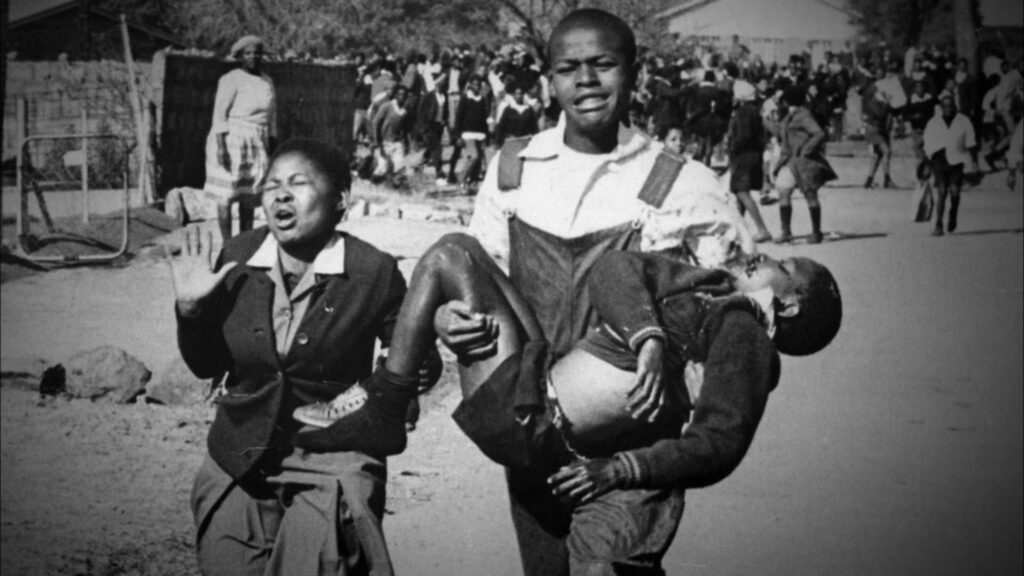 Black and white historical photograph by Sam Nzima of the 1976 Soweto Uprising. A distressed young man carries the body of Hector Pieterson, while a girl runs alongside them in grief.